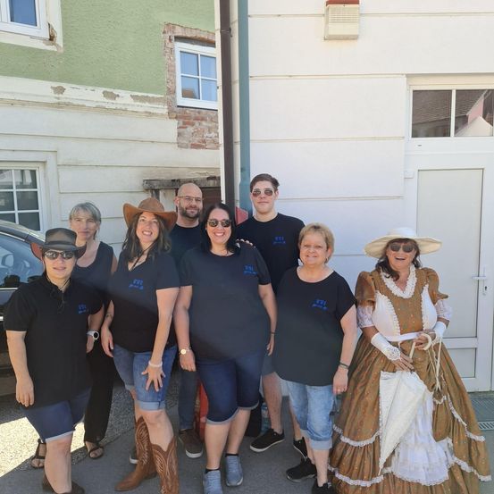 Eine Gruppe von Menschen in verschiedenen Outfits steht vor einem Gebäude. Einige tragen Cowboyhüte, andere viktorianische Kleider. Sie alle lächeln für die Kamera.