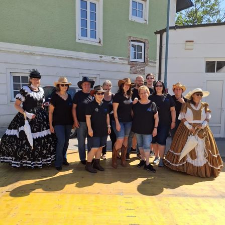 Eine Gruppe von Menschen in 19. Jahrhundert Kleidung steht vor einem Gebäude. Sie lächeln und scheinen für ein Foto zu posieren. Hinter ihnen sind zwei weiße Gebäude mit Fenstern.