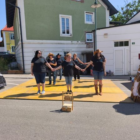 Eine Gruppe von Frauen in schwarzen T-Shirts hält sich an einem gelben Podest in einem Parkplatz die Hände. Sie stehen vor einem grünen Gebäude.