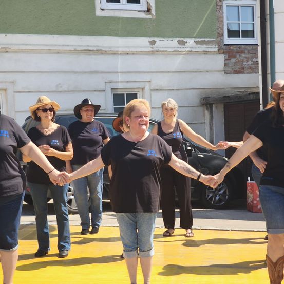 Eine Gruppe von Menschen steht im Kreis und hält sich an den Händen vor einem Gebäude. Sie tragen Hüte und Sonnenbrillen. Ein Auto ist hinter ihnen geparkt.