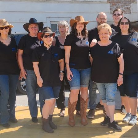 Eine Gruppe von Menschen in schwarzen T-Shirts und Jeansshorts, einige mit Cowboy-Hüten, stehen draußen in der Nähe eines Autos und lächeln für ein Foto.