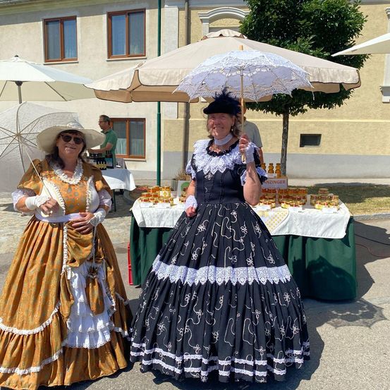 Zwei Frauen in traditioneller Kleidung stehen unter Regenschirmen im Freien. Eine hält einen weißen Schirm, und beide lächeln für die Kamera. Dahinter steht ein Tisch mit Gläsern und ein Gebäude mit Fenstern.