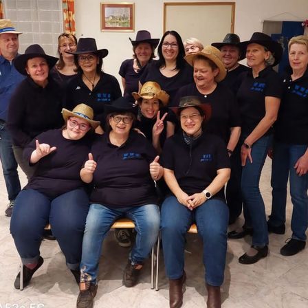 Eine Gruppe von Frauen in Cowboy-Huten posiert für ein Foto. Sie tragen alle passende schwarze T-Shirts. Einige stehen, andere sitzen auf einer Bank.