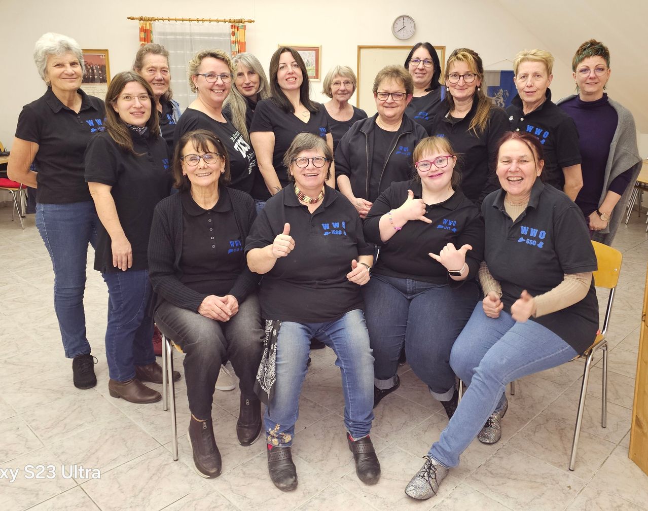 Eine Gruppe von Frauen in gleichen schwarzen T-Shirts mit blauer Schrift posiert für ein Foto, einige mit Brille und Daumen hoch. Sie sitzen auf Stühlen in einem Raum mit gefliestem Boden.