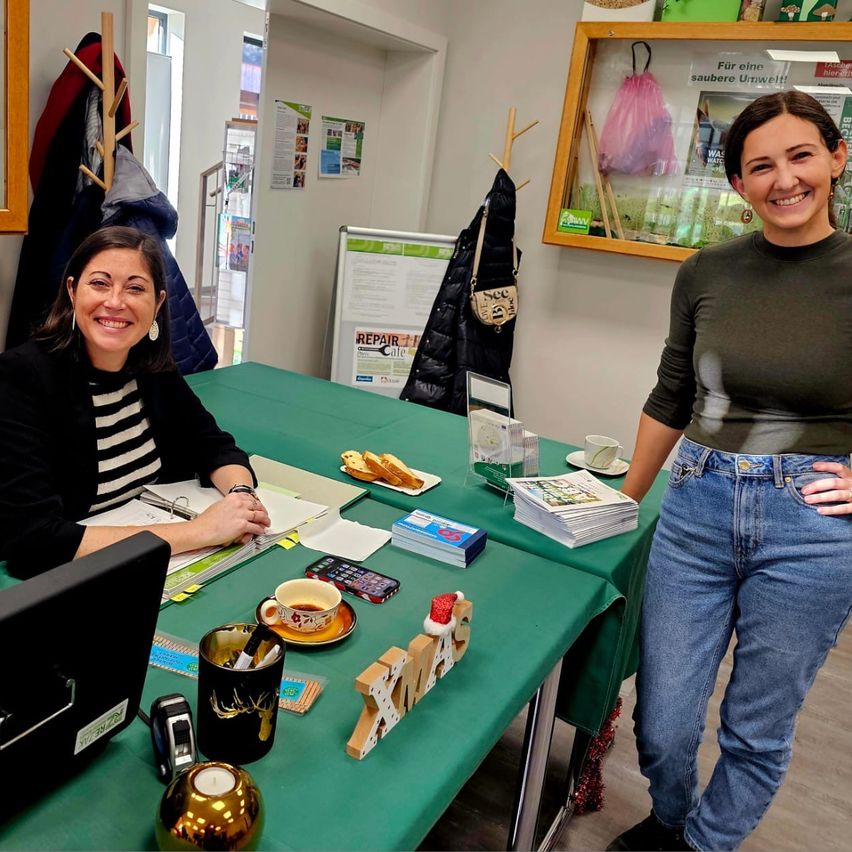 Zwei Frauen stehen bei einem Tisch mit Weihnachtsdekoration und Essen, eine lächelt. Hinter ihnen hängen Kleidung an einem Ständer und ein Bulletin-Board zeigt einen Aushang.