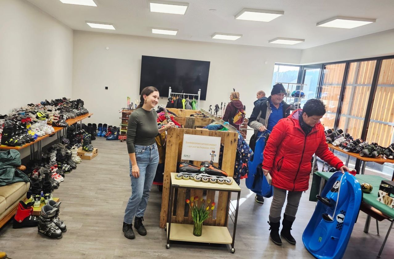 Eine Frau steht mit einem Lächeln in einem Geschäft, umgeben von Regalen mit Schuhen und Kleidung. Eine andere Frau in einer roten Jacke steht in der Nähe und hält einen Gegenstand. Ein Mann in einem blauen Shirt und schwarzem Beanie ist ebenfalls anwesend.