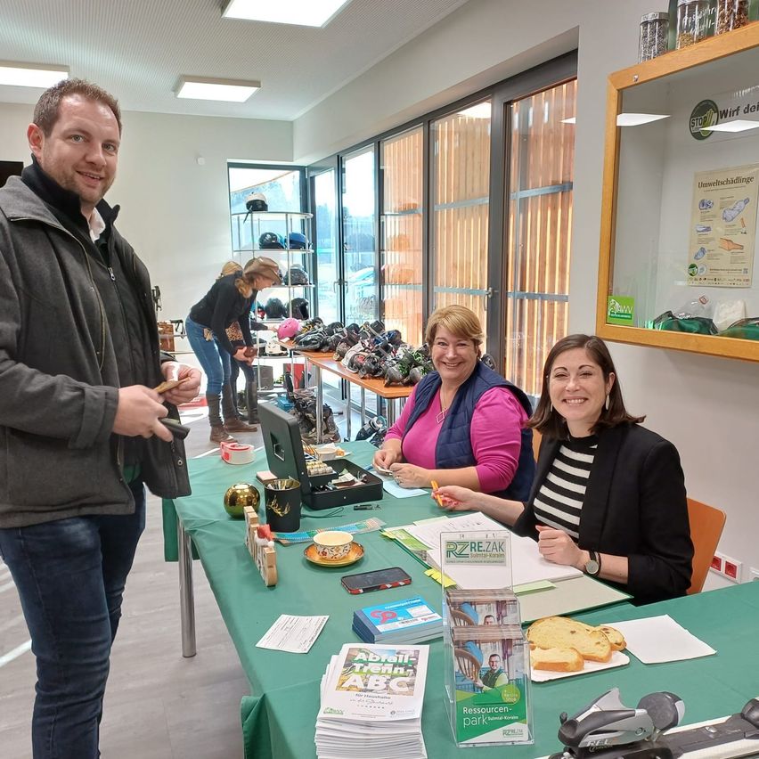 Drei Personen, zwei Frauen und ein Mann, stehen hinter einem Tisch mit verschiedenen Gegenständen. Eine Frau hält einen Stift, während eine andere Frau lächelt. Ein Regal mit verschiedenen Gegenständen ist im Hintergrund zu sehen.