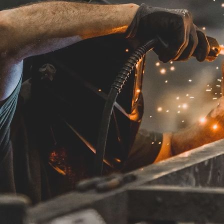 Eine Person in einer Schweißmaske hält eine Schweißfackel und arbeitet mit Funkenflug an einem Metallstück. Die Person trägt Handschuhe und ein grünes Shirt.