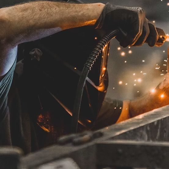 Eine Person in einer Schweißmaske hält eine Schweißfackel und arbeitet mit Funkenflug an einem Metallstück. Die Person trägt Handschuhe und ein grünes Shirt.