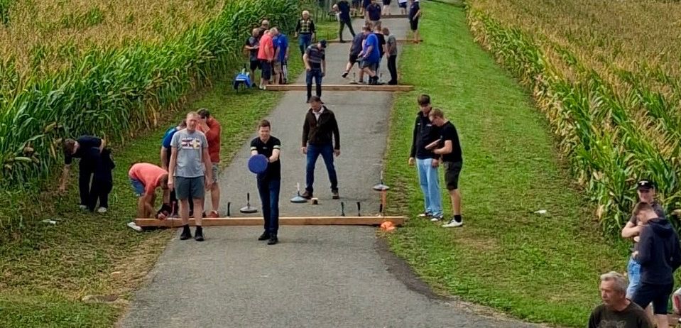 Eine Gruppe von Menschen versammelt sich auf einem Weg in einem Maisfeld, einige spielen ein Spiel, andere stehen und beobachten. Autos sind am Straßenrand geparkt.