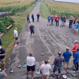 Eine Gruppe von Menschen ist auf einer Straße versammelt, möglicherweise für ein Spiel. Einige tragen Freizeitkleidung, während andere Mützen und Turnschuhe tragen. Einige Leute stehen in der Nähe der Straße, während andere auf dem Gras stehen. Einige Gegenstände sind auf der Straße platziert. In der Ferne gibt es ein Feld mit Mais und ein paar Autos.