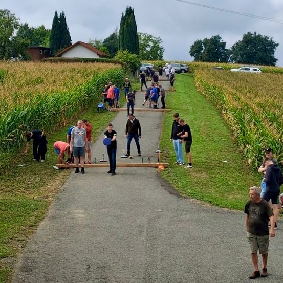 Eine Gruppe von Menschen spielt ein Spiel auf einem Pfad in einem Maisfeld, einige beugen sich über und andere stehen herum. Ein Haus und geparkte Autos sind im Hintergrund zu sehen.