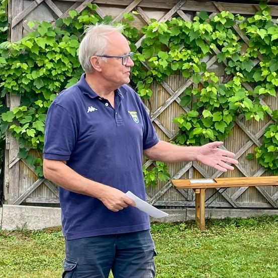 Ein älterer Mann in einem blauen Poloshirt steht im Freien und gestikuliert mit einer Hand, während er ein Stück Papier in der anderen hält. Hinter ihm befindet sich ein mit grünen Reben bedeckter Holzzaun.