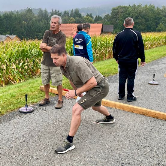 Mehrere Männer spielen ein Spiel in einem Maisfeld, wobei sich ein Mann in Sneakers bückt. Ein Mann in einem schwarzen Mantel beobachtet von hinten.