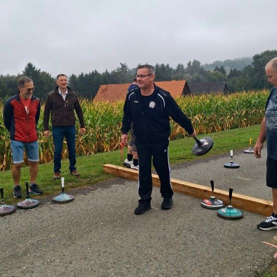 Fünf Männer stehen auf einem Schotterfeld und spielen möglicherweise ein Spiel. Ein Mann hält einen Schläger, und ein anderer Mann steht in der Nähe und beobachtet. Dahinter befindet sich ein Maisfeld und Häuser in der Ferne.