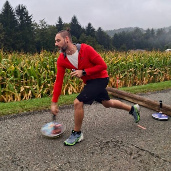 Ein Mann in einer roten Kapuzenjacke läuft auf einem Pfad in einem Maisfeld, hält einen Frisbee und eine Wasserflasche.