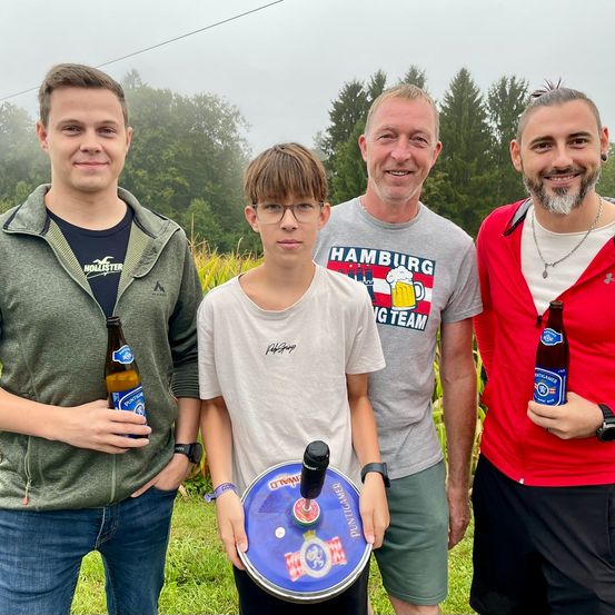 Vier Männer stehen lächelnd in einem Feld, zwei halten Bierflaschen und ein Junge einen Frisbee. Sie tragen Freizeitkleidung und stehen vor Bäumen.