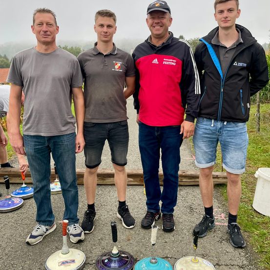 Vier Männer stehen zusammen im Freien. Sie tragen Freizeitkleidung, einschließlich Sneaker und Poloshirts. Ein Mann trägt einen Hut mit dem Wort 'Feiting'. Sie stehen auf einem Betonuntergrund mit verschiedenen Objekten vor ihnen. Hinter ihnen befindet sich eine grasbewachsene Fläche und ein Holzzaun.