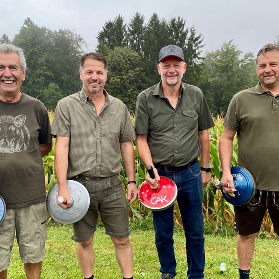 Vier Männer stehen in einem Grasfeld, lächeln und posieren für ein Foto. Sie tragen alle Freizeitkleidung und halten blaue und rote Scheiben. Bäume und Mais sind im Hintergrund.