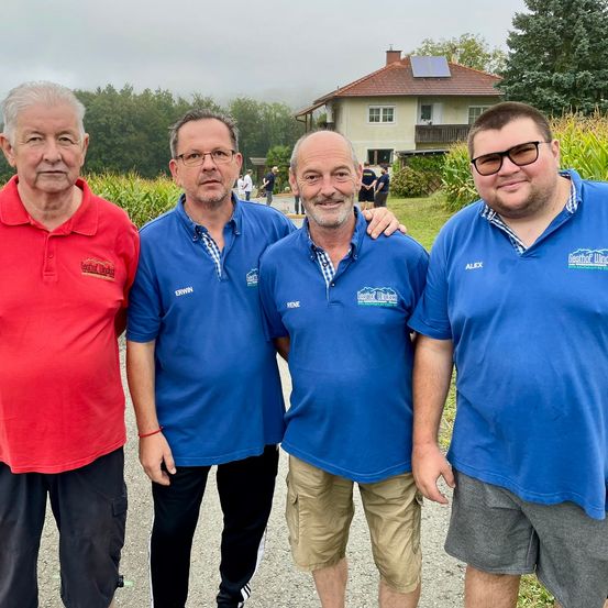 Vier Männer stehen lächelnd auf einem Pfad vor einem Haus. Drei sind in blauen Shirts und einer in rot. Dahinter befindet sich ein Feld mit Mais und ein Haus mit Solarpanelen.