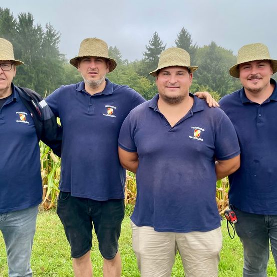 Vier Männer stehen in einem Grasfeld mit Bäumen im Hintergrund. Sie tragen blaue Polo-Shirts mit Logos, Strohhüte und einer hat eine Kamera. Der Hintergrund zeigt einen Wald mit dichten Bäumen.
