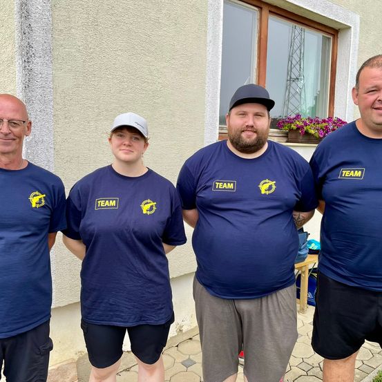 Vier Personen stehen vor einem Gebäude und lächeln für ein Foto. Sie tragen marineblaue T-Shirts mit gelben Team-Logos. Der Mann rechts trägt einen Hut, und eine Frau links trägt eine weiße Kappe. Hinter ihnen befindet sich ein Fenster mit einem Blumentopf.