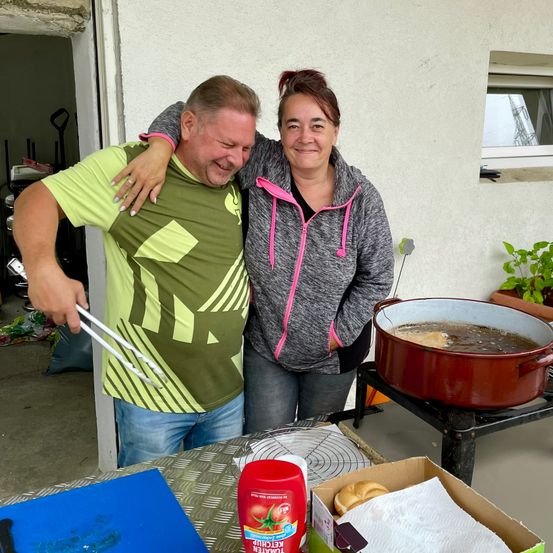 Ein Mann und eine Frau lächeln und posieren für ein Foto in einer Küche. Sie stehen neben einem Topf mit einer Pfanne auf dem Herd. Auf dem Tisch stehen eine Pflanze, eine Flasche und eine Box. Hinter ihnen befindet sich eine Wand mit einem Fenster und einer Tür.