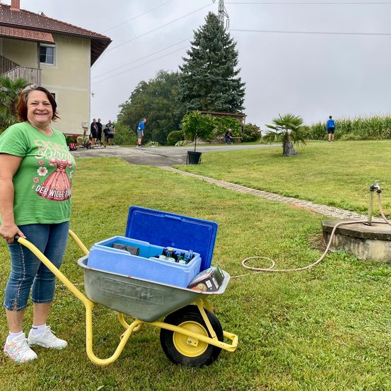 Eine Frau schiebt einen Schubkarren mit einem blauen Kühler voller Getränke auf einem Rasen. Hinter ihr steht ein Haus mit Menschen, die gehen und stehen. Bäume, Pflanzen und ein Weg sind ebenfalls vorhanden.