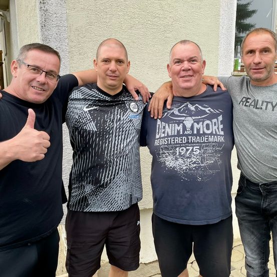 Vier Männer posieren für ein Foto vor einem Gebäude. Der Mann auf der linken Seite gibt einen Daumen hoch. Der Mann in der Mitte trägt ein Shirt mit einem Bergdesign und dem Text 'Denim and More'. Der Mann auf der rechten Seite trägt ein graues Shirt mit dem Wort 'REALITY'.