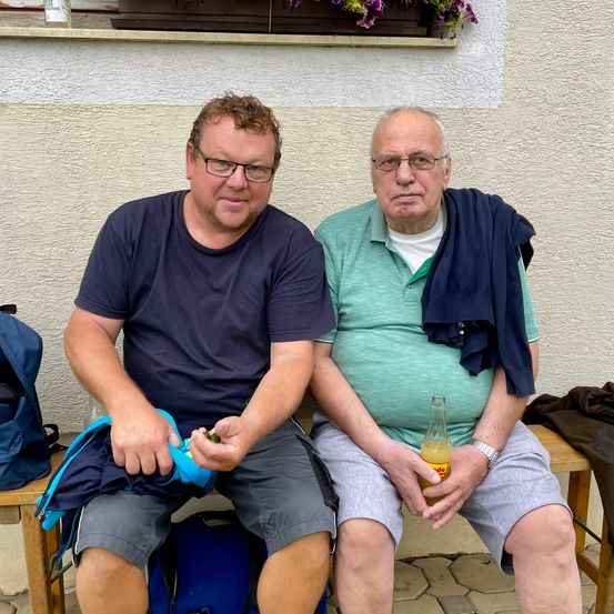 Zwei Männer sitzen auf einer Bank. Einer hat eine blaue Jacke über der Schulter, der andere hält eine Flasche. Ein Rucksack liegt in der Nähe.