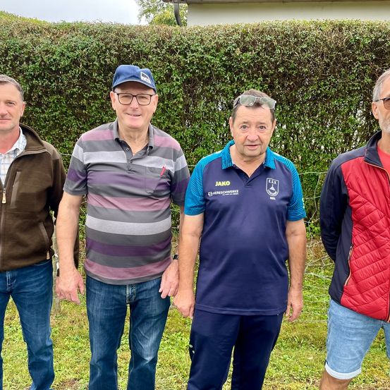 Vier Männer stehen vor einer Hecke. Der Mann auf der linken Seite trägt eine braune Jacke. Der Mann neben ihm trägt ein gestreiftes Hemd. Der dritte Mann trägt ein blaues Poloshirt mit dem Wort 'Jako' darauf. Der Mann auf der rechten Seite trägt eine rot-schwarze Jacke.