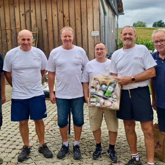 Fünf Männer stehen zusammen auf einem Kiesweg. Sie tragen weiße T-Shirts und Shorts, und einer hält einen Korb mit Essen. Sie lächeln und scheinen für ein Foto zu posieren. Hinter ihnen steht ein Holzgebäude und ein Feld.