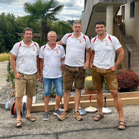 Vier Männer in weißen Poloshirts mit roten Einsätzen und braunen Shorts stehen zusammen vor einem Haus mit einer Treppe und Pflanzen.