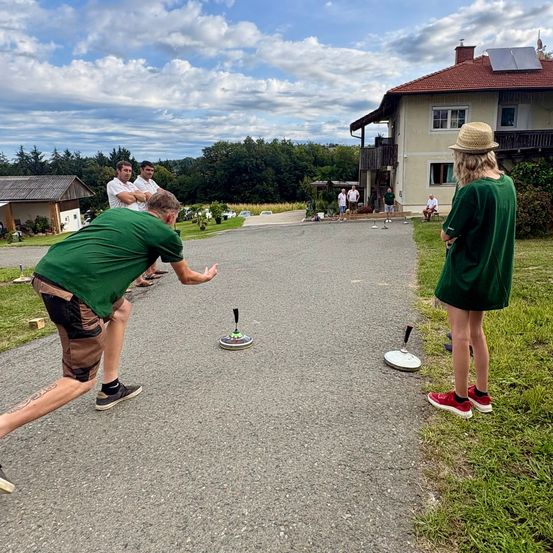 Eine Gruppe von Menschen spielt ein Cornhole-Spiel auf einer Einfahrt vor einem Haus mit Solarpanelen auf dem Dach.