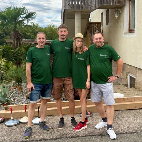Vier Personen in grünen T-Shirts stehen vor einem Haus mit Garten und lächeln für ein Foto. Eine Frau trägt einen Hut.