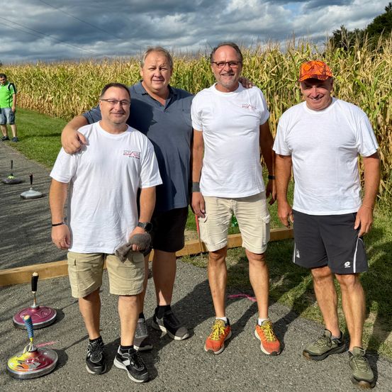 Vier Männer stehen vor einem Maisfeld und lächeln für ein Foto. Sie tragen Freizeitkleidung und Turnschuhe. Einer hält ein Handtuch, und hinter ihnen sind Maispflanzen und ein Grasbereich.