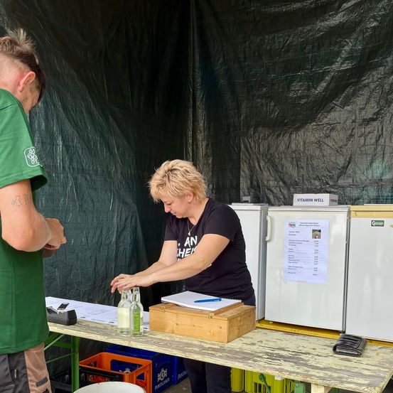 Eine Frau in einem schwarzen T-Shirt füllt ein Getränk in eine Flasche an einem Tisch mit einer Box und einem Blatt Papier. Ein Mann in einem grünen T-Shirt steht hinter ihr.