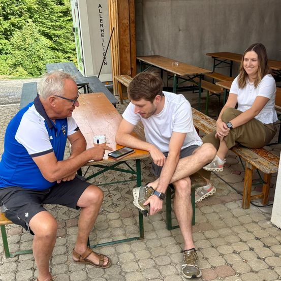 Drei Personen sitzen auf Bänken im Freien, ein Mann schaut auf sein Handy, während ein anderer Mann ihn ansieht. Eine Frau lächelt im Hintergrund.