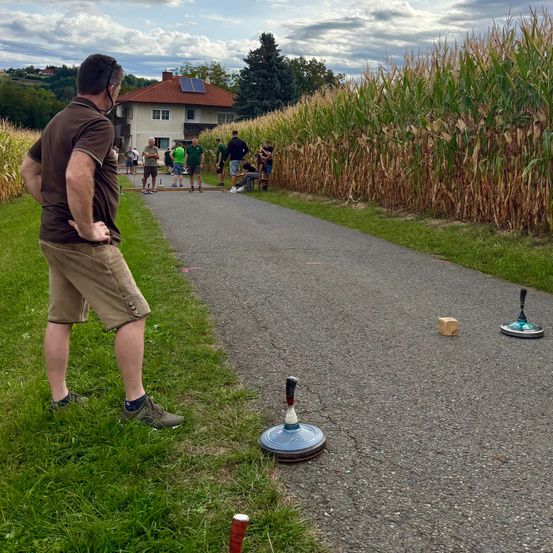 Ein Mann steht auf einem Pfad in einem Maisfeld und bereitet sich darauf vor, einen Ball auf ein Ziel zu werfen. In der Nähe versammeln sich Leute und schauen zu.
