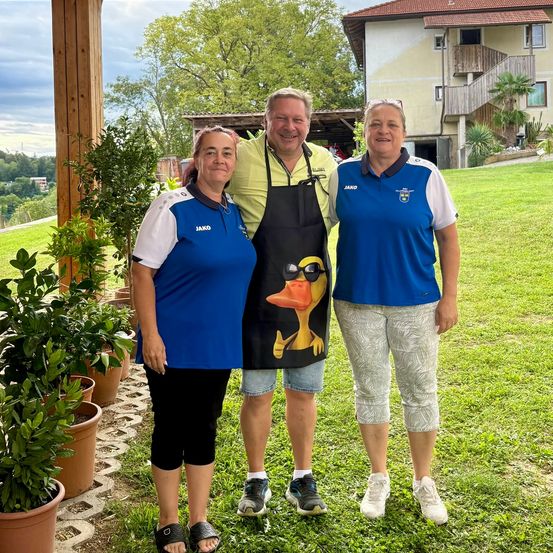 Drei Personen stehen auf einem Rasen vor einem Haus mit einer Treppe und einem Balkon. Zwei Frauen tragen blaue und weiße Uniformen, und ein Mann trägt ein gelbes Hemd und eine Schürze mit Entendesign.