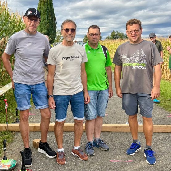 Vier Männer stehen zusammen in einem Maisfeld und lächeln für ein Foto. Sie tragen lässige Sommerkleidung, einschließlich Mützen und Turnschuhe. Dahinter sind eine Holzstruktur und ein Grasbereich sichtbar.