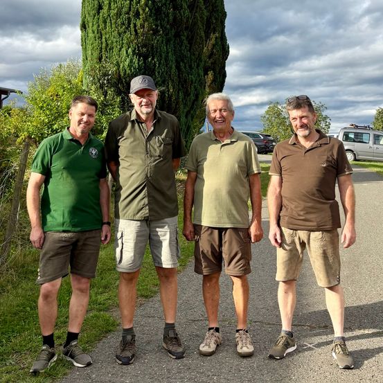 Vier Männer stehen auf einem Kiesweg und lächeln für ein Foto. Sie tragen lässige Outdoor-Kleidung und stehen vor einem hohen Baum und einem bewölkten Himmel.