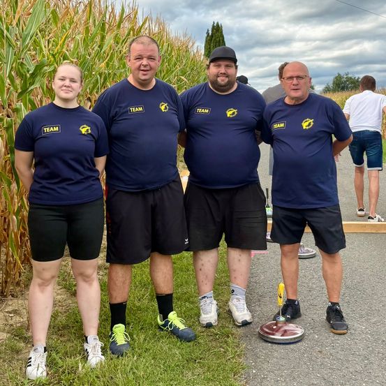 Vier Personen in passenden blauen T-Shirts mit dem Wort 'Team' darauf stehen vor einem Maisfeld. Sie lächeln und posieren für ein Foto.