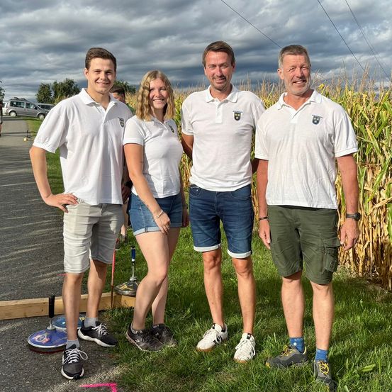 Vier Personen in weißen Poloshirts und lässigen Shorts stehen auf einem Rasenfeld, lächeln und posieren für ein Foto. Hinter ihnen befindet sich ein Maisfeld und ein bewölkter Himmel.
