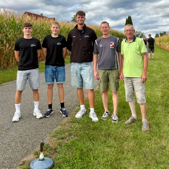 Fünf Männer stehen in einer Reihe auf einem Weg und lächeln für ein Foto. Sie tragen Freizeitkleidung und Turnschuhe. Hinter ihnen befindet sich ein Grasbereich mit einem Maisfeld und einigen Menschen, die spazieren gehen.