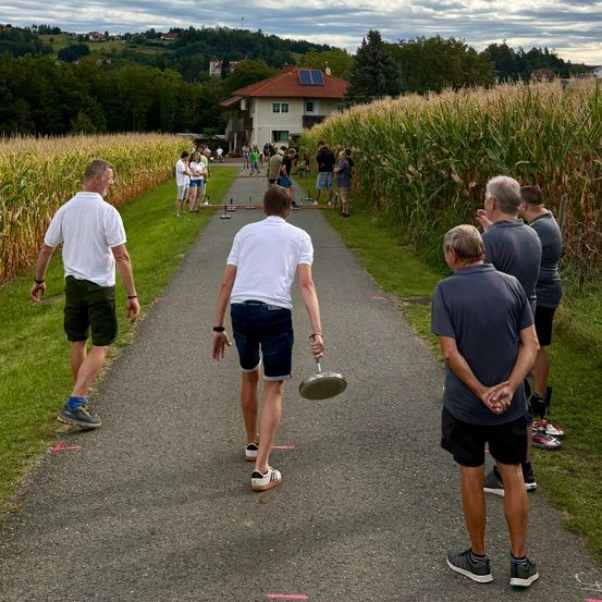 Eine Gruppe von Männern spielt Discgolf auf einem Pfad, umgeben von hohem Mais und einem Haus mit Solarpanelen.