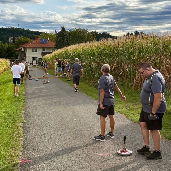 Eine Gruppe von Menschen spielt eine Partie Kegel auf einem Betonweg, umgeben von einem Maisfeld und einem Haus mit Solarmodulen.
