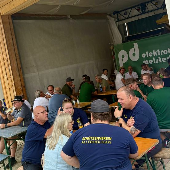 Eine Gruppe von Menschen in blauen und grünen Shirts sitzt an Tischen in einem Zelt, einige unterhalten sich und andere trinken. Im Hintergrund ist ein grünes Banner mit weißem Text zu sehen.