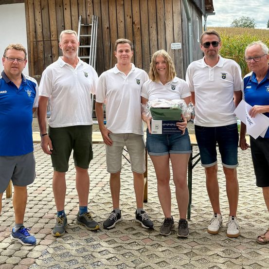 Fünf Personen stehen vor einem Holzgebäude und lächeln für ein Foto. Die Frau in der Mitte hält eine Geschenkbox. Der Mann auf der linken Seite trägt ein blaues Shirt, die anderen weiße Hemden.