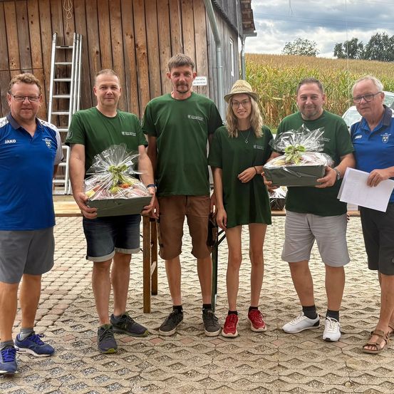 Eine Gruppe von Menschen in grünen Shirts und Shorts steht vor einem Holzgebäude und hält Geschenkkörbe. Sie lächeln und posieren für ein Foto.